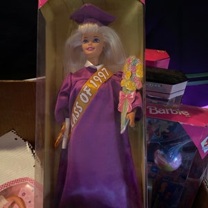Puede incluir: Una muñeca Barbie con un vestido de graduación morado y un gorro. Tiene una banda dorada que dice "Clase de 1997". Lleva un diploma y un ramo de flores. La caja dice "Barbie de graduación edición especial".