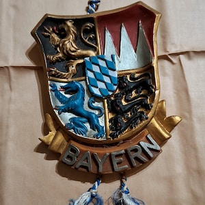 Puede incluir: Un escudo decorativo con la palabra "BAYERN" en letras plateadas. El escudo está dividido en cuadrantes con símbolos heráldicos, incluyendo leones y un patrón de cuadros azules y blancos. Tiene un borde dorado y una cuerda azul y blanca para colgar.
