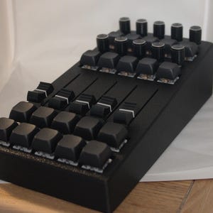 May include: A black macro pad featuring a grid of square keys and four vertical sliders on the front section, with a row of eight rotary encoder knobs positioned at the top. The device has a matte black finish and a rectangular, angled housing.
