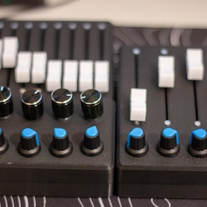 Puede incluir: Primer plano de dos superficies de control de audio negras con deslizadores y perillas blancas. Las perillas son negras con tapas plateadas y detalles azules. Los deslizadores son blancos. Para producción musical o mezcla de audio.