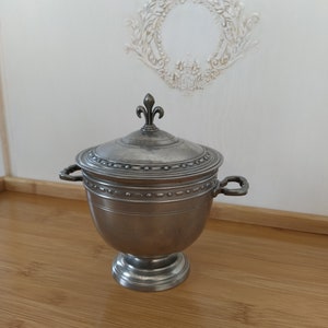 May include: A silver pewter sugar bowl with a lid and two handles. The lid has a fleur-de-lis finial.