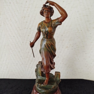 May include: A bronze-coloured statue of a woman in a flowing dress, holding a stick and raising her arm. The statue is on a multi-tiered base with a reddish-brown wooden finish. The woman's dress has green and brown tones.