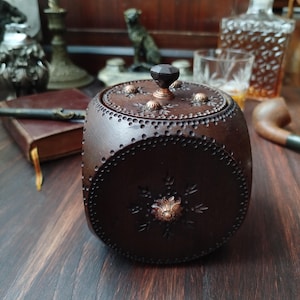 Peut inclure: Une boîte ronde en bois brun foncé avec un couvercle. La boîte est ornée de petits accents ronds de couleur laiton et d'un motif floral central. Le couvercle a un bouton sombre à facettes. La boîte est probablement destinée à ranger de petits objets.