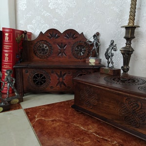 Può includere: Una panca in legno scuro intagliata con motivi intricati e una scatola di legno abbinata. La panca ha uno schienale rialzato e un pannello frontale intagliato. La scatola ha un coperchio incernierato e un top intagliato. Entrambi i pezzi sono su un pavimento piastrellato.