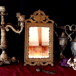 May include: Ornate gold-colored framed mirror with a decorative top and base. The mirror is rectangular and stands on a red velvet surface. Other antique items, including candlesticks and vases, are visible in the background.