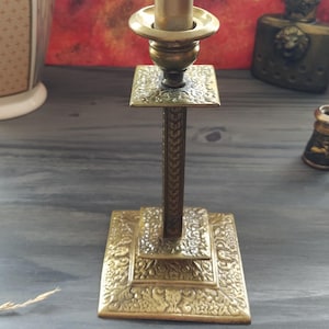 May include: A gold metal candlestick with a square base and a twisted candle holder. The base is decorated with an intricate floral design.