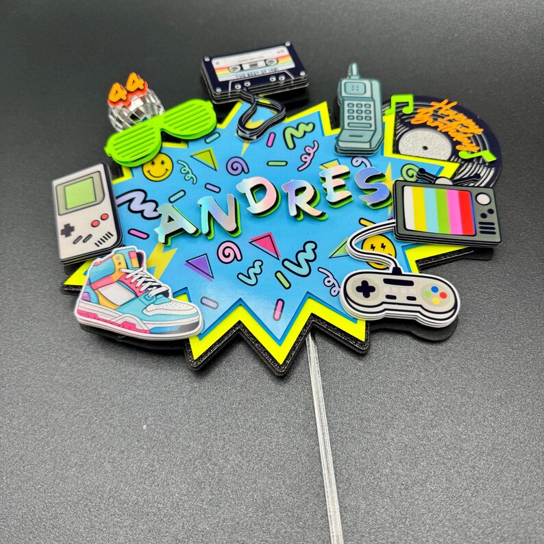 May include: A cake topper featuring a blue starburst shape with the name ANDRES in iridescent letters. It is decorated with 1980s and 1990s themed icons including a cassette tape, handheld game console, high-top sneaker, phone, and controller.
