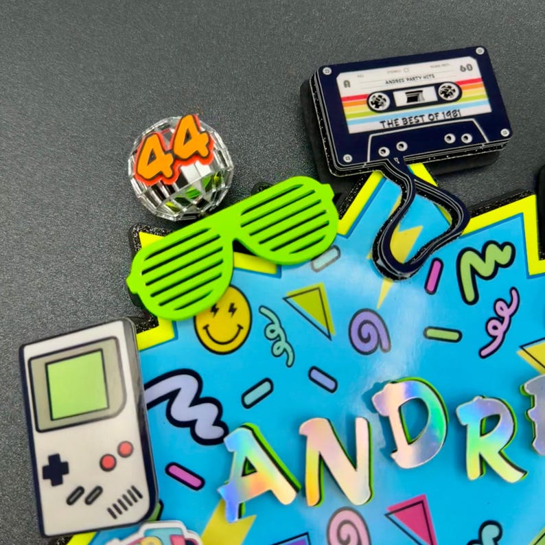 May include: A decorative cake topper featuring a blue background with colorful geometric shapes, a white handheld game console, a silver disco ball with the number 44, neon green shutter glasses, and a black cassette tape labeled The Best of 1981.