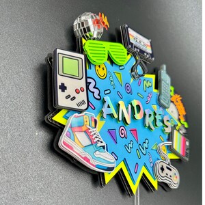 May include: A multi-layered cake topper featuring the name ANDRES in iridescent letters on a blue starburst background. It includes 1980s-themed icons: a handheld game console, high-top sneaker, disco ball, shutter shades, cassette tape, and controller.