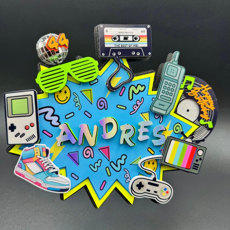May include: A multi-layered cake topper featuring a blue starburst background with the name ANDRES in iridescent letters. It is decorated with 1980s-themed cutouts including a disco ball with 44, cassette tape, shutter glasses, brick phone, sneaker, game console, and TV.