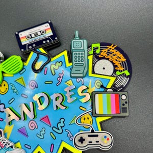 May include: A decorative cake topper featuring a bright blue starburst background with the name ANDRES in holographic letters. It includes 1980s-themed icons: a cassette tape, a brick cell phone, a vinyl record, a television, and a game controller.