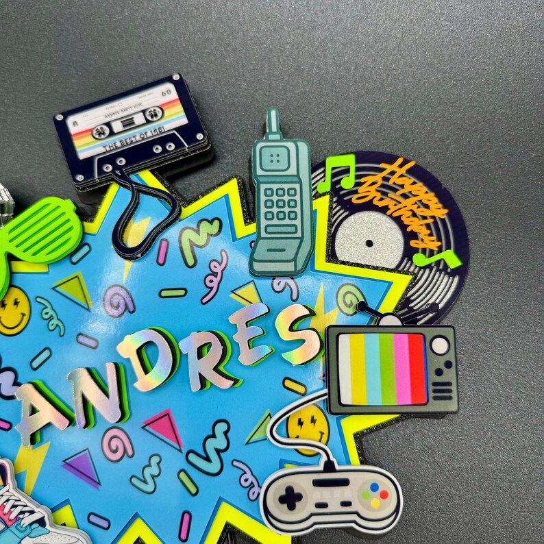 May include: A decorative cake topper featuring a bright blue starburst background with the name ANDRES in holographic letters. It includes 1980s-themed icons: a cassette tape, a brick cell phone, a vinyl record, a television, and a game controller.