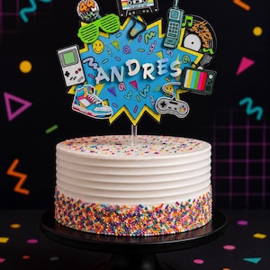 May include: A white frosted cake with horizontal ridges and colorful sprinkles at the base, topped with a blue starburst cutout featuring the name ANDRES and 1980s-themed icons including a cassette tape, game controller, sneaker, and disco ball.