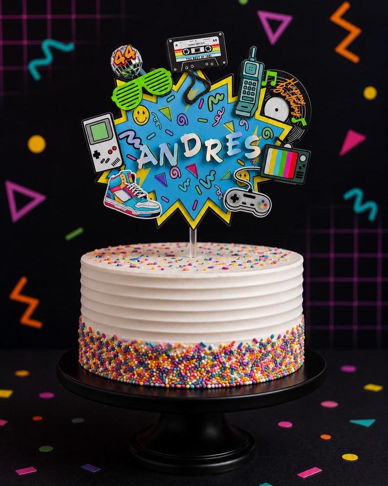 May include: A white frosted cake with horizontal ridges and colorful sprinkles at the base, topped with a blue starburst cutout featuring the name ANDRES and 1980s-themed icons including a cassette tape, game controller, sneaker, and disco ball.