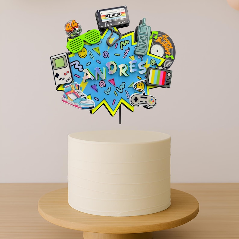 May include: A cake topper featuring a blue starburst shape with the name ANDRES in the center, surrounded by 1990s-themed icons including a cassette tape, handheld game console, high-top sneaker, mobile phone, television, and game controller.