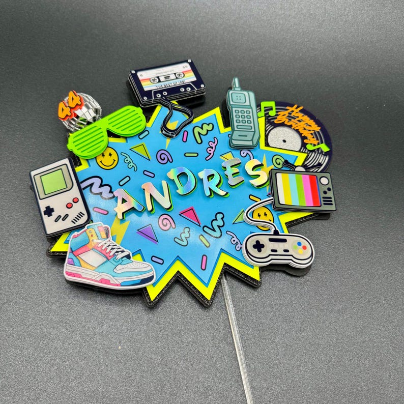 May include: A cake topper featuring a blue starburst shape with the name ANDRES in the center. It is decorated with 1990s-themed icons including a handheld game console, a cassette tape, a mobile phone, a sneaker, a television, and a game controller.