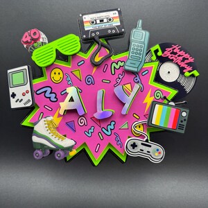May include: A decorative cake topper featuring the name ALY in iridescent letters on a pink starburst background. It is surrounded by 1980s-themed icons including a cassette tape labeled ALY'S PARTY HITS, a handheld game console, a roller skate, a mobile phone, a vinyl record with the text Happy Birthday, a television, and a game controller.