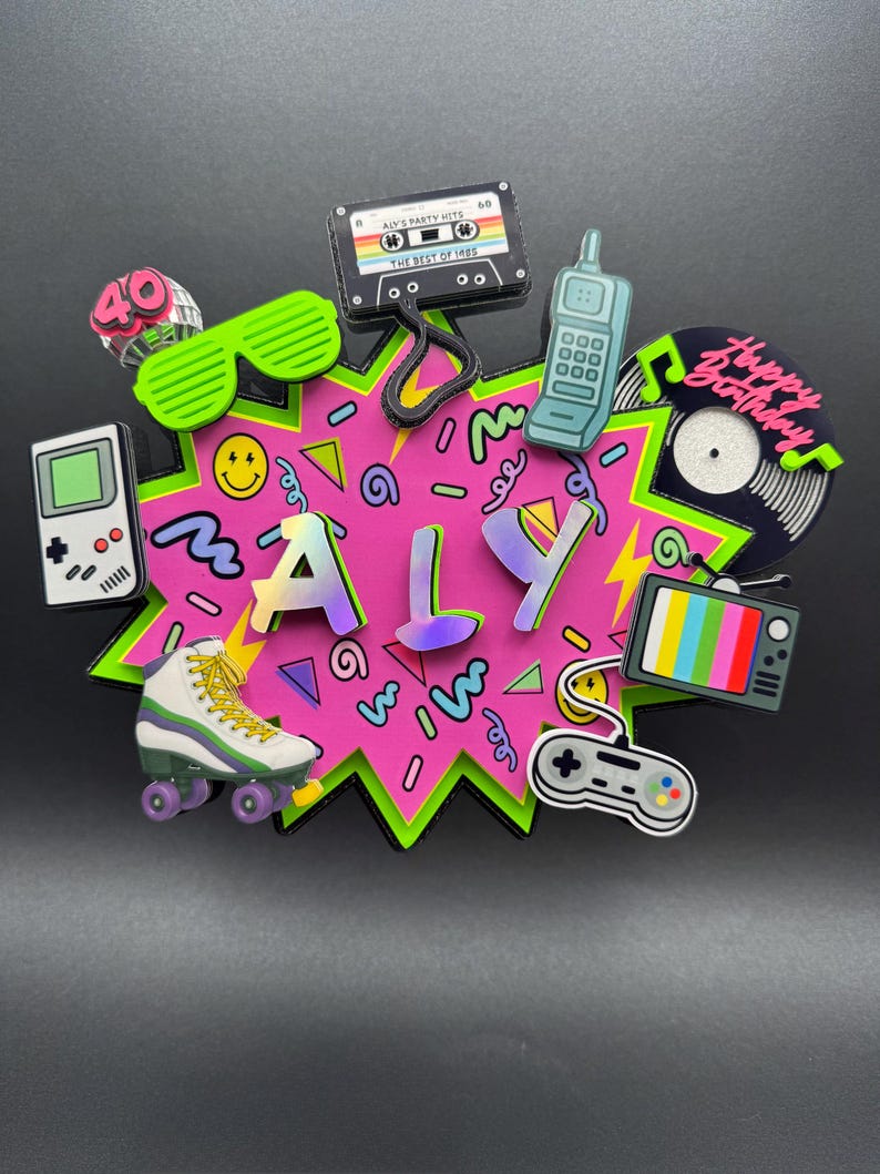 May include: A decorative cake topper featuring the name ALY in iridescent letters on a pink starburst background. It is surrounded by 1980s-themed icons including a cassette tape labeled ALY'S PARTY HITS, a handheld game console, a roller skate, a mobile phone, a vinyl record with the text Happy Birthday, a television, and a game controller.
