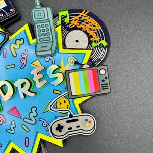May include: A blue star-shaped cake topper featuring a retro theme with a teal brick cell phone, a vinyl record with the text Happy Birthday, a television with color bars, and a gray video game controller. The topper has holographic lettering.
