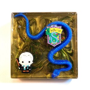 以下が含まれることがあります： 青いヘビ、杖を持った小さなキャラクター、緑と青の蛇の家の紋章が描かれた正方形の樹脂製コースター。背景は緑と赤の渦巻き模様の金色です。