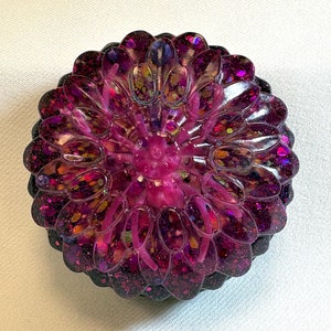 Peut inclure: Une fleur ronde en résine rose et violette avec des paillettes incrustées dans la résine. La fleur a un aspect multicouche et dimensionnel.