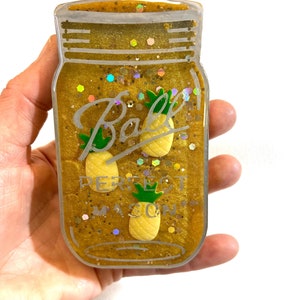 May include: A gold mason jar with glitter and two yellow pineapple charms. The jar has the text "Ball Perfect Mason" on it.