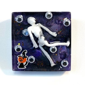 May include: A square resin paperweight with a purple and black background. A white skeleton is floating in the resin with glitter and googly eyes. A small pumpkin with a witch's hat is in the bottom left corner.