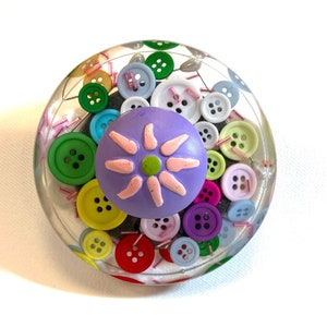 May include: A clear, round paperweight with a colorful assortment of buttons embedded inside. The buttons are various sizes and colors, including green, yellow, red, and blue. A purple flower-shaped embellishment sits on top, adding a whimsical touch.