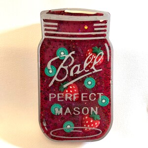 以下が含まれることがあります： 銀色の蓋と「Ball Perfect Mason」という文字が書かれた赤いガラス瓶。瓶は赤い液体で満たされ、緑と赤の小さなプラスチック製の果物で飾られています。