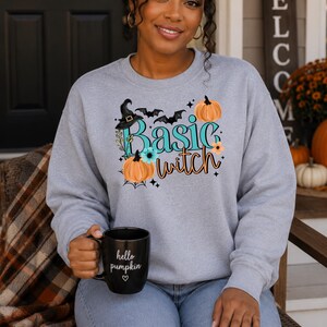 May include: A light gray crewneck sweatshirt featuring a graphic print that says Basic witch in teal and orange text. The design includes a black witch hat, orange pumpkins, small bats, and flowers. A black mug with the text hello pumpkin is held.