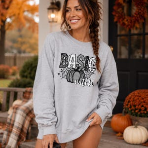 May include: A light gray crewneck sweatshirt featuring a black graphic print on the chest. The design includes the word BASIC in block letters, a central pumpkin illustration, spider web patterns, and the word witch in cursive script.