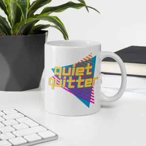 Könnte beinhalten: Weiße Keramiktasse mit einem geometrischen Design in Blau, Rosa und Gelb. Der Text "Quiet Quitter" ist in gelben Buchstaben geschrieben.