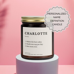 Puede incluir: Una vela de vidrio marrón con una tapa dorada. La etiqueta dice "CHARLOTTE (nombre) 1. Hace los mejores pasteles. 2. Necesita una siesta al día. 3. La mejor mamá de todas. 9 oz | 50 horas de tiempo de combustión | 100% vegano". La vela está sobre una superficie de color púrpura claro. Una forma de estrella blanca con el texto "VELA DE DEFINICIÓN DE NOMBRE PERSONALIZADA" está en la esquina superior derecha.