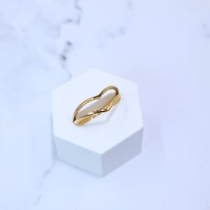 May include: A gold-colored ring with a heart-shaped design. The ring is displayed on a white hexagonal stand against a white and gray marble background. The ring's design features a curved band that forms a heart shape.