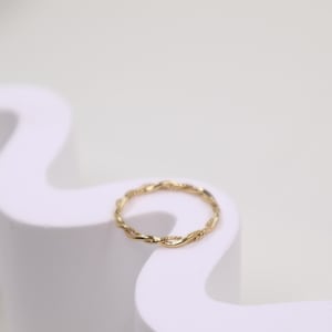 May include: A delicate gold ring with a twisted rope design. The ring is set against a white background, highlighting its intricate details. This piece of jewellery is a simple yet elegant accessory.