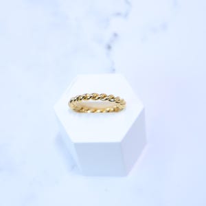 May include: A gold ring featuring a twisted rope design. The ring is presented on a white hexagonal stand against a white and light blue backdrop. The ring's design is a continuous, textured pattern.