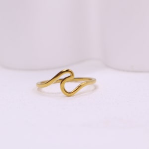 May include: A gold-coloured ring with a wave design. The ring features a continuous, flowing line that forms a stylized wave shape. The ring is set against a white background, highlighting its simple yet elegant design. This piece of jewellery is suitable for everyday wear.