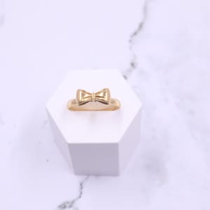 May include: A gold-coloured ring featuring a bow design. The ring is displayed on a white hexagonal stand.