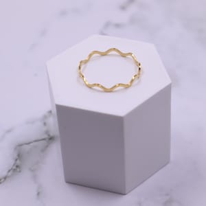 May include: A gold-coloured ring with a wavy design. The ring is on a white hexagonal display stand.
