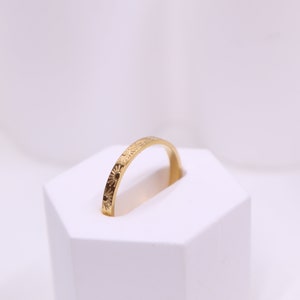 May include: A gold ring with a detailed floral pattern. The ring is displayed on a white hexagonal stand against a soft, white background. The ring's design features small, repeating sunburst and floral motifs.