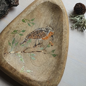 May include: Wooden, free-form tray with a hand-painted bird perched on a branch, holding berries. The bird has brown and orange feathers. Green leaves and gold accents decorate the tray. A piece of bark and a pine cone are in the background.
