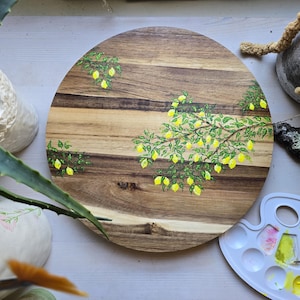 Puede incluir: Una bandeja redonda de madera para servir con un diseño pintado de una rama con hojas verdes y limones amarillos. La bandeja tiene un acabado de veta de madera natural.