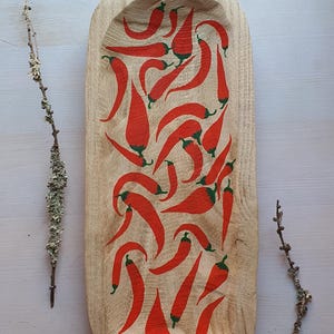 Puede incluir: Bandeja de madera oblonga con un centro tallado, decorada con diseños de pimientos rojos. Los pimientos tienen tallos verdes. La bandeja es de color marrón claro y descansa sobre una superficie blanca. Ramitas con líquenes en los lados.