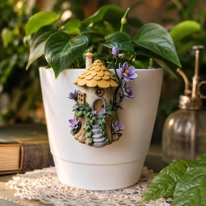 May include: A white ceramic plant pot with a decorative fairy house attached. The fairy house has a yellow roof, a small door, and purple flowers. Green leaves of a plant are visible.