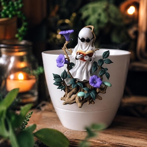 May include: A white ceramic planter with a whimsical ghost figurine reading a book. The ghost wears sunglasses and is surrounded by purple flowers and green leaves. The planter is set against a blurred background with a candle.