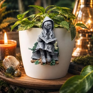Puede incluir: Una maceta de cerámica blanca con una estatua gris detallada de una persona leyendo un libro, con una capa con capucha y gafas. La maceta contiene una planta de hojas verdes. Una vela encendida y otras decoraciones están en el fondo.