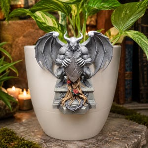 May include: A cream-colored plant pot with a gray gargoyle statue attached. The statue has a horned head, large wings, and is holding a shield. Red roses and a chain are at the base of the statue.