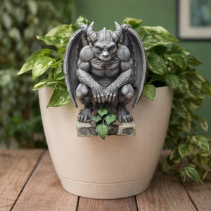 Puede incluir: Una estatua de gárgola gris con cuernos y alas, colocada frente a una maceta beige llena de plantas verdes y blancas. La gárgola está sobre una base de piedra con hojas verdes. La estatua está en cuclillas.