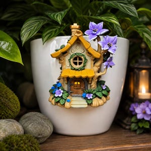 May include: A white ceramic planter with a whimsical fairy house attached. The house has a yellow roof, a brown door, and is adorned with purple flowers and green foliage. A decorative accent for indoor or outdoor use.