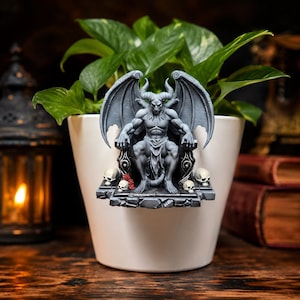 May include: A gray, winged, horned demon figurine seated on a throne, attached to the rim of a white planter. The base of the throne features small skulls and red roses. The demon has a muscular build, pointed ears, and a tail, rendered in a matte finish.